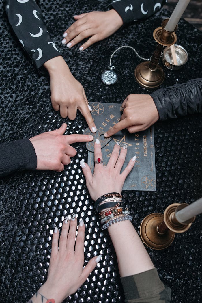 Four diverse hands using a Ouija board with candles on a dark textured surface.